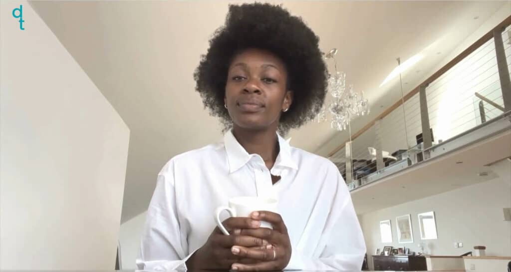 A black deaf woman sits down at a glass table. She holds her coffee cup on the table whilst talking. Her background is an open plan room with bright lighting. She wears a baggy white blouse. She speaks in English. There is a black deaf woman, who wears a red long-sleeved shirt with black pants, in the right corner, who is translating the spoken English to British Sign Language.
