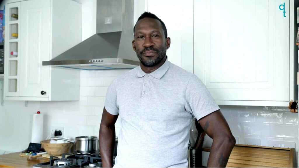 A black deaf man stands in his kitchen. The cabinets are all white and there is a silver cheese grater directly behind him. There are various kitchen items on the table behind him. He wears a grey polo shirt. He signs in British Sign Language.