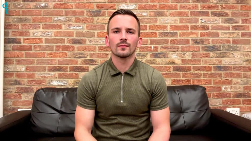 A white deaf man sits on a black leather sofa. His background is a brick wall which is a mixture of red, brown and cream. He wears a khaki polo shirt with a three-quarter zip. He signs in British Sign Language.